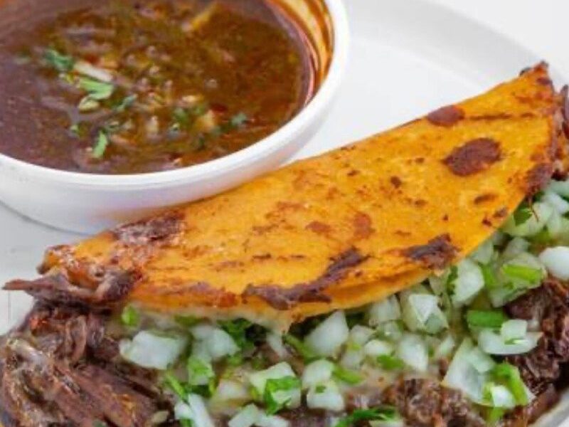 Birria taco with onions and cilantro next to a cup of consomé